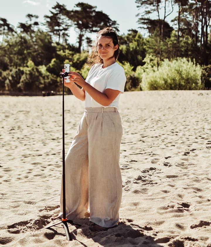 Mobile video maker i content creator Agata Dargacz w trakcie pracy. Zdjęcie w stylu "behind the scenes" pokazuje Agatę na słonecznej, piaszczystej plaży, gdy ustawia smartfon na statywie, przygotowując się do nagrywania wideo w plenerze. To idealny przykład nowoczesnego tworzenia autentycznych treści na social media.